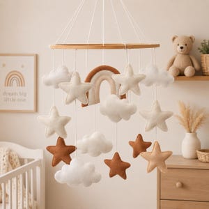May include: A baby mobile featuring felt stars, clouds, and a rainbow in neutral tones. The mobile hangs from a wooden ring and is suspended by white strings. A framed print with the text "dream big little one" is visible.