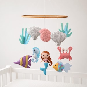 May include: A baby mobile featuring felt sea creatures and shells. The mobile includes a mermaid, seahorse, crab, and fish, along with glittery shells and coral. The wooden ring suspends the colorful ocean-themed decorations.