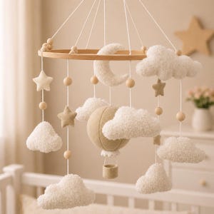 Puede incluir: Móvil de bebé artesanal con un aro de madera, nubes de fieltro, estrellas y una luna creciente. Un globo aerostático beige cuelga en el centro. El móvil está diseñado con colores neutros.