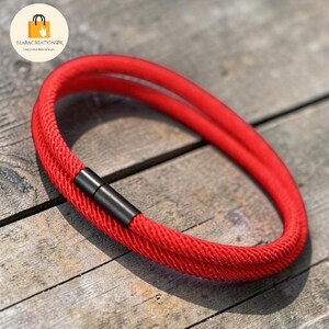 May include: A vibrant red bracelet with a braided texture and a black magnetic clasp. The bracelet is circular and appears to be made of a thick, durable material. The background is a weathered wooden surface.