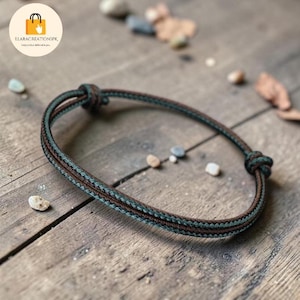 May include: A handmade bracelet with a braided design. The bracelet features a combination of brown and green threads, knotted at both ends. The bracelet is displayed on a wooden surface, with small stones and leaves scattered around.