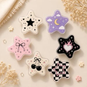 Puede incluir: Seis recipientes con forma de estrella con diseños distintos: una estrella blanca con una estrella negra, una estrella morada con luna y nubes, una estrella rosa con un lazo, una estrella negra con un corazón en llamas, una estrella blanca con cerezas y una estrella a cuadros blancos y negros.