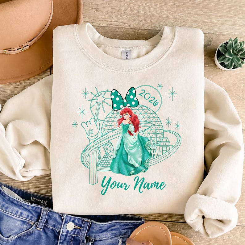 Pu&ograve; includere: Felpa color crema con grafica turchese di una principessa con capelli rossi, fiocco a pois e la scritta "2020". Il design include un parco a tema e le parole "Your Name".