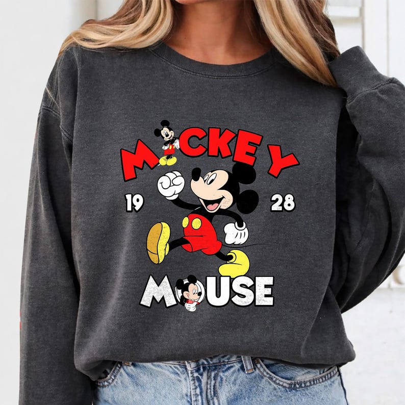 Puede incluir: Sudadera gris oscuro con un dise&ntilde;o de Mickey Mouse. El dise&ntilde;o incluye la palabra "Mickey" en rojo, el a&ntilde;o "1928" y la palabra "Mouse" en blanco. Mickey lleva pantalones cortos rojos y zapatos amarillos.