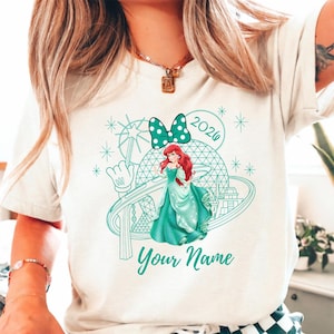 Pu&ograve; includere: T-shirt color crema con grafica turchese di una principessa in abito turchese, un fiocco a pois e la scritta "Your Name" sotto. La grafica include anche l'anno "2026" e una mano di Topolino.