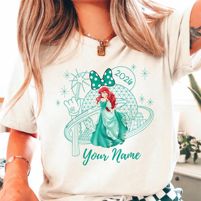 Pu&ograve; includere: T-shirt color crema con grafica turchese di una principessa in abito turchese, un fiocco a pois e la scritta "Your Name" sotto. La grafica include anche l'anno "2026" e una mano di Topolino.