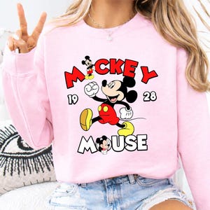 Puede incluir: Sudadera rosa claro con un dise&ntilde;o de Mickey Mouse. El dise&ntilde;o incluye la palabra "Mickey" en rojo, "Mouse" en blanco y los n&uacute;meros "19" y "28". Mickey se representa en su pose ic&oacute;nica.