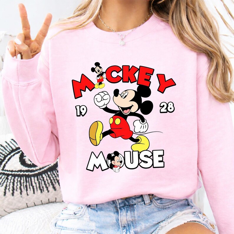 Puede incluir: Sudadera rosa claro con un dise&ntilde;o de Mickey Mouse. El dise&ntilde;o incluye la palabra "Mickey" en rojo, "Mouse" en blanco y los n&uacute;meros "19" y "28". Mickey se representa en su pose ic&oacute;nica.