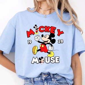 Puede incluir: Camiseta azul claro con un dise&ntilde;o de Mickey Mouse en movimiento. El dise&ntilde;o incluye las palabras "Mickey Mouse" en rojo y blanco, junto con los n&uacute;meros "19" y "28". Un estilo cl&aacute;sico y casual.
