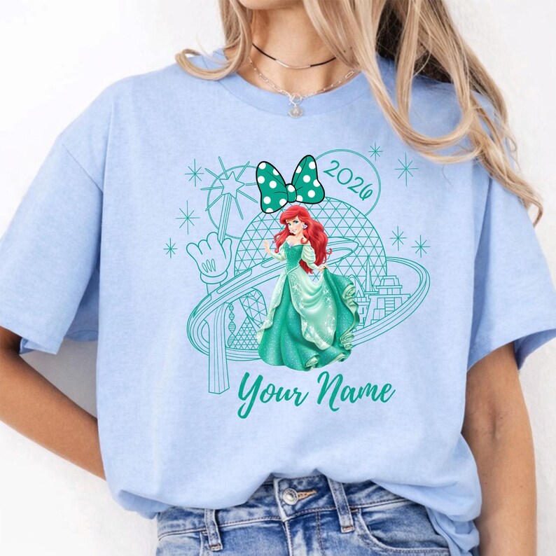Pu&ograve; includere: T-shirt azzurra con grafica verde acqua di una principessa in abito verde, fiocco a pois e l'anno 2020. Il testo "Your Name" &egrave; sotto la principessa. La maglietta &egrave; in stile casual.