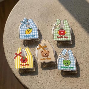 May include: Five small, bag-shaped phone grips featuring colorful patterns and characters. One is blue plaid with a lemon and 'LEMON DAY', one green plaid with an apple and 'Apple day', one yellow striped with a tomato and 'TOMATO', one beige with a bear and 'nice day', and one blue plaid with a green fruit and 'SUNNY DAY'.