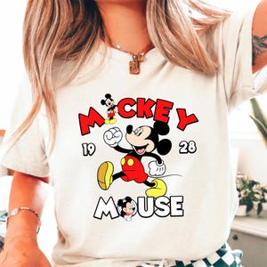 Puede incluir: Camiseta color crema con un dise&ntilde;o de Mickey Mouse. El dise&ntilde;o incluye la palabra "Mickey" en rojo, el a&ntilde;o "1928" y la palabra "Mouse" en contorno blanco. Mickey Mouse aparece con pantalones cortos rojos y zapatos amarillos.