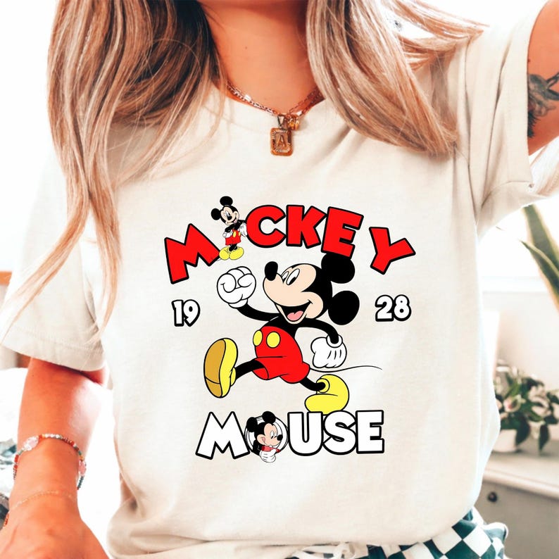 Puede incluir: Camiseta color crema con un dise&ntilde;o de Mickey Mouse. El dise&ntilde;o incluye la palabra "Mickey" en rojo, el a&ntilde;o "1928" y la palabra "Mouse" en contorno blanco. Mickey Mouse aparece con pantalones cortos rojos y zapatos amarillos.