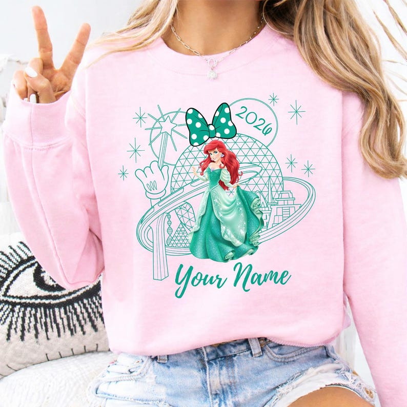 Pu&ograve; includere: Felpa rosa chiaro con grafica turchese di una principessa, fiocco a pois e scritta "2026". Il design include una ruota panoramica e la scritta "Your Name".
