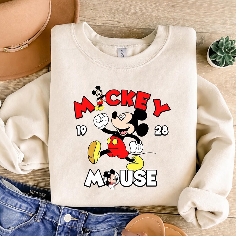 Puede incluir: Una sudadera crema con un dise&ntilde;o de Mickey Mouse en rojo, negro, amarillo y blanco. El dise&ntilde;o incluye la palabra "Mickey" en rojo, el a&ntilde;o "1928" y la palabra "Mouse" en blanco. La sudadera est&aacute; doblada sobre una superficie de madera.