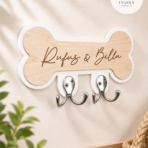 May include: A bone-shaped wall hook rack featuring a light wood finish with a white border. The text "Rufus & Bella" is engraved in a dark script font across the centre. Two polished silver metal double hooks are attached to the bottom edge.