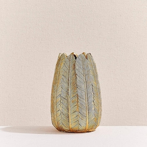 Puede incluir: Un jarrón decorativo con diseño de plumas. El jarrón es principalmente dorado y verde, con una superficie texturizada. El jarrón tiene una abertura ancha y una base redondeada. El fondo es de color neutro.
