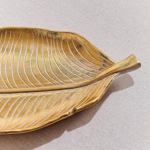 Puede incluir: Una bandeja decorativa dorada en forma de hoja con una superficie texturizada y detalles blancos. La bandeja tiene un patrón de vetas detallado y una forma ligeramente curvada, ideal para guardar objetos pequeños. La bandeja es de metal.