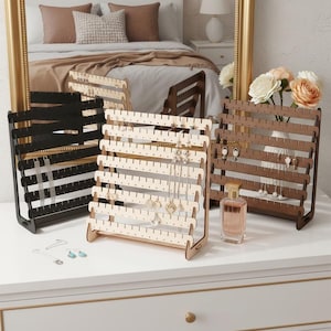 Peut inclure: Trois présentoirs à bijoux à plusieurs niveaux en finitions noir, bois clair et bois foncé. Chaque support comporte plusieurs barres horizontales avec des encoches régulièrement espacées pour suspendre des boucles d'oreilles et des colliers.