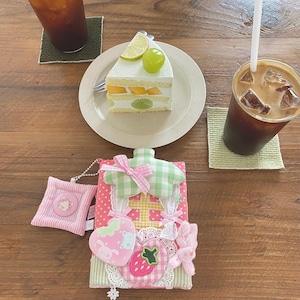 Könnte beinhalten: Ein Stück mehrschichtiger Kuchen mit Obstbelag, eine rosa-weiß gemusterte Hülle mit Erdbeer- und Hasenmuster und zwei Eiskaffee-Getränke auf einem Holztisch. Der Kuchen hat Mango- und Kiwischeiben.