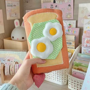 Puede incluir: Una funda de cuaderno de tela diseñada para parecerse a una rebanada de pan tostado con dos huevos fritos. La funda tiene un patrón de cuadros amarillos y naranjas, con una forma hexagonal verde con lunares. Los huevos tienen yemas amarillas y claras blancas.