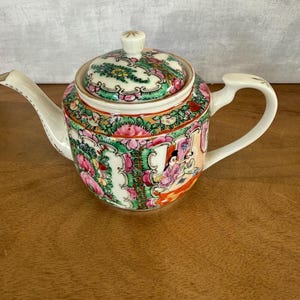 Puede incluir: Una tetera de porcelana con tapa, con un diseño floral y figurativo colorido. La tetera tiene un asa y un pico blancos, y la tapa está rematada con un pequeño pomo. El diseño incluye flores rosas, verdes y amarillas, y una escena de personas.
