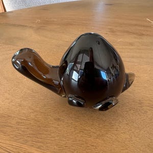 Op de afbeelding: Een donkerbruin glazen schildpadbeeldje met een glanzende afwerking. De schildpad heeft een afgeronde schild, een kop en vier kleine pootjes. Het beeldje staat op een houten oppervlak.