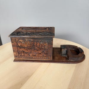 Könnte beinhalten: Ein dunkelbrauner Schreibtisch-Organizer aus Holz mit einer rechteckigen Box, die auf der Vorder- und Oberseite mit detaillierten Blumenmustern verziert ist. An der Seite der Box befindet sich ein runder Halter für kleine Gegenstände.