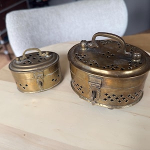 Op de afbeelding: Twee ovale koperen doosjes met scharnierende deksels en gebogen handvatten. Elke container heeft een geperforeerd patroon van kleine kruisjes aan de zijkanten en op het deksel. De doosjes hebben een sluiting aan de voorzijde en een verweerde goudkleurige afwerking.