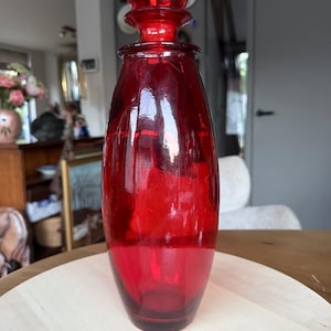 May include: A tall, translucent red glass decanter with a smooth, rounded body and a matching spherical glass stopper. The vessel features a narrow neck that widens towards the centre before tapering slightly at the base, showcasing a vibrant crimson hue.