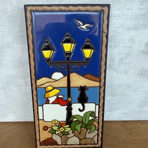 Op de afbeelding: Een rechthoekige keramische tegel met een nachtscène met een zwarte lantaarnpaal, een persoon met een gele hoed en een zwarte kat op een witte muur. De achtergrond toont een blauwe lucht, een witte maan, een vliegende vogel en bruine heuvels.