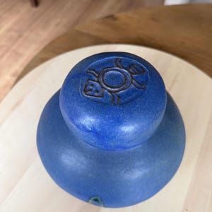 Può includere: Un coperchio in ceramica blu cobalto con una maniglia a pomello arrotondata. La parte superiore del pomello presenta un grafico stampato con contorni scuri di una tartaruga stilizzata. La superficie della ceramica ha una finitura opaca e testurizzata.