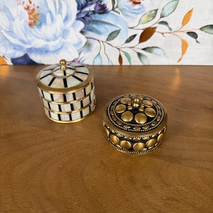 Op de afbeelding: Twee kleine ronde decoratieve doosjes. Het eerste doosje heeft een wit en zwart gestreept patroon met een messing rand en een kleine messing knop op het deksel. Het tweede doosje is van donker metaal met verhoogde goudkleurige cirkels.