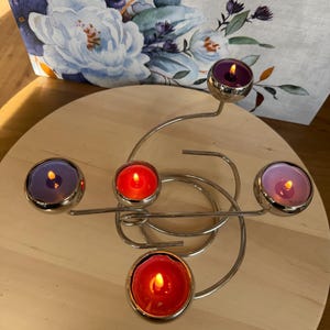 Pode incluir: Um suporte de vela em metal prateado com uma base em espiral e quatro copos esféricos. Cada copo contém uma pequena vela de chá com uma chama acesa. As velas são de tons violeta, vermelho e rosa claro, criando um brilho quente.