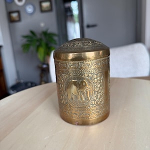 Op de afbeelding: Een cilindrische koperen container met een afneembaar deksel, voorzien van ingewikkelde reliëfpatronen en een centraal olifantenmotief. Het metalen oppervlak vertoont een warme, gepolijste goudtint met gedetailleerde gravures rondom de olifant.