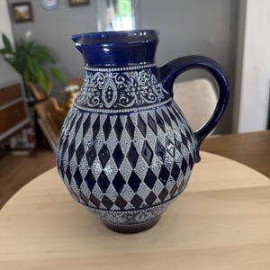 Może przedstawiać: Ceramiczny dzbanek z ciemnoniebieskim szkliwem na krawędzi i uchwycie, z korpusem ozdobionym powtarzającym się wzorem rombów w kolorze ciemnoniebieskim i teksturowanym jasnoszarym. Szyjka i podstawa są ozdobione misternymi białymi tłoczeniami.