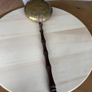 May include: A brass skimmer with a perforated circular head and a long, dark brown wooden handle featuring a bamboo-like texture. The handle terminates in a small metal loop at the end, and the brass head is attached to the handle with metal rivets.