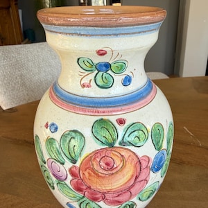 Puede incluir: Un jarrón de cerámica pintado a mano con base color crema. El jarrón presenta una gran rosa central en tonos rojos y naranjas, rodeada de hojas verdes y detalles azules y rosas. El borde es marrón.