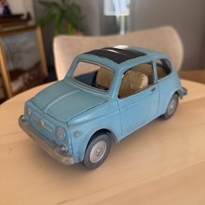 Könnte beinhalten: Ein hellblaues Spielzeugauto aus Metall mit schwarzem Schiebedach, beigen Innensitzen und silberfarbenen Rädern. Das Fahrzeug hat eine abgerundete Karosserieform mit einer vorderen Stoßstange, runden Scheinwerfern und einem kleinen Emblem auf der Motorhaube.