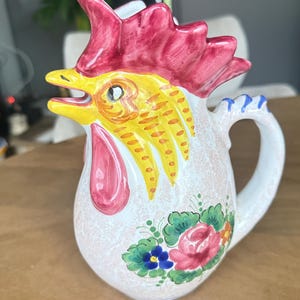 Può includere: Una brocca in ceramica a forma di gallo con una cresta rosso vivo, testa gialla e bargigli rosa. Il corpo è bianco sporco con una consistenza maculata e presenta un motivo floreale dipinto a mano nei toni del rosa, verde e blu.