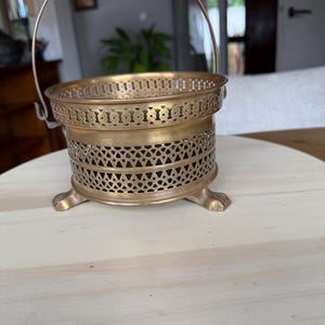 Puede incluir: Una cesta redonda de latón con un intrincado patrón de celosía calada en los laterales y un asa metálica fina y arqueada. El recipiente se apoya sobre tres pequeñas patas en forma de garra y tiene un acabado metálico pulido con pátina.
