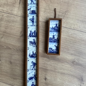 Op de afbeelding: Twee verticale wandhangers met blauw-witte keramische tegels in houten lijsten. De lange lijst bevat negen tegels met figuren en landschappen, terwijl de kortere lijst drie tegels met afbeeldingen van windmolens bevat.