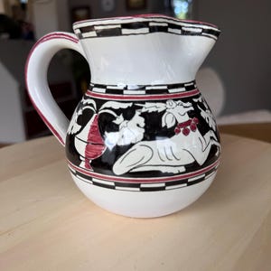 Pode incluir: Um jarro de cerâmica branca com um design pintado à mão em preto e vermelho. O padrão inclui uma borda xadrez ao redor do gargalo e da base, com uma faixa central que retrata um cervo deitado, folhagem estilizada e um motivo de vaso.