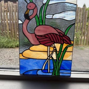Op de afbeelding: Een rechthoekig glas-in-loodpaneel met een roze flamingo die in blauw water staat, met geel zand en groene rietstengels. De achtergrond bestaat uit grijze en witte glassegmenten. Het geheel is samengevoegd door zwarte loden lijnen.