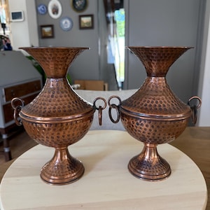 Könnte beinhalten: Ein Paar passende Kupfervasen mit gehämmertem Muster, einem weit ausgestellten Rand, einem bauchigen Körper und einem Standfuß. Jede Vase ist mit zwei kleinen, runden Metallringgriffen an den Seiten des Hauptkörpers verziert.