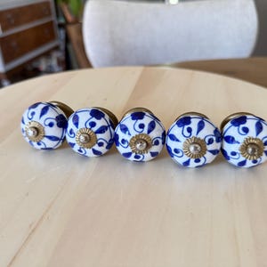 Może przedstawiać: Pięć okrągłych ceramicznych gałek do szafek z białą podstawą, niebieskim wzorem kwiatowym i centralnym akcentem z metalu w kolorze mosiądzu. Każda gałka ma gładkie, błyszczące wykończenie i okrągły kształt, przeznaczony do montażu na meblach.