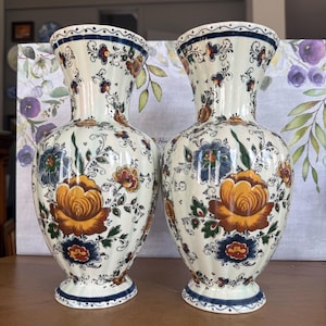 Op de afbeelding: Twee bijpassende keramische vazen met een crèmekleurige basis en bloempatronen. De vazen hebben een grote gele roos, blauwe en oranje bloemen en groene bladeren. De randen en bases zijn blauw afgewerkt. De vazen staan op een houten oppervlak.