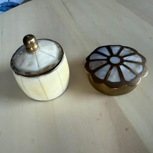 May include: A cylindrical container with a cream-colored, vertically striped lid and a matching base, paired with a round, scalloped-edge brass box featuring a white, petal-like inlay pattern on its lid. Both items have metallic brass accents.