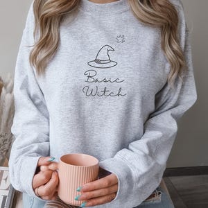 Könnte beinhalten: Ein hellgraues Sweatshirt mit Rundhalsausschnitt und einer schwarzen Strichzeichnung eines Hexenhutes sowie eines kleinen Sterns. Darunter befindet sich der Schriftzug "Basic Witch" in Schreibschrift. Das Oberteil hat lange Ärmel und Rippbündchen.