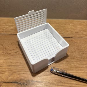 Könnte beinhalten: Ein weißer Schreibtisch-Organizer mit Klappdeckel und einem Stapel liniertes Papier. Der Organizer hat einen geschwungenen Ausschnitt auf einer Seite. Ein schwarzer und blauer Stift liegt auf der Holzoberfläche.